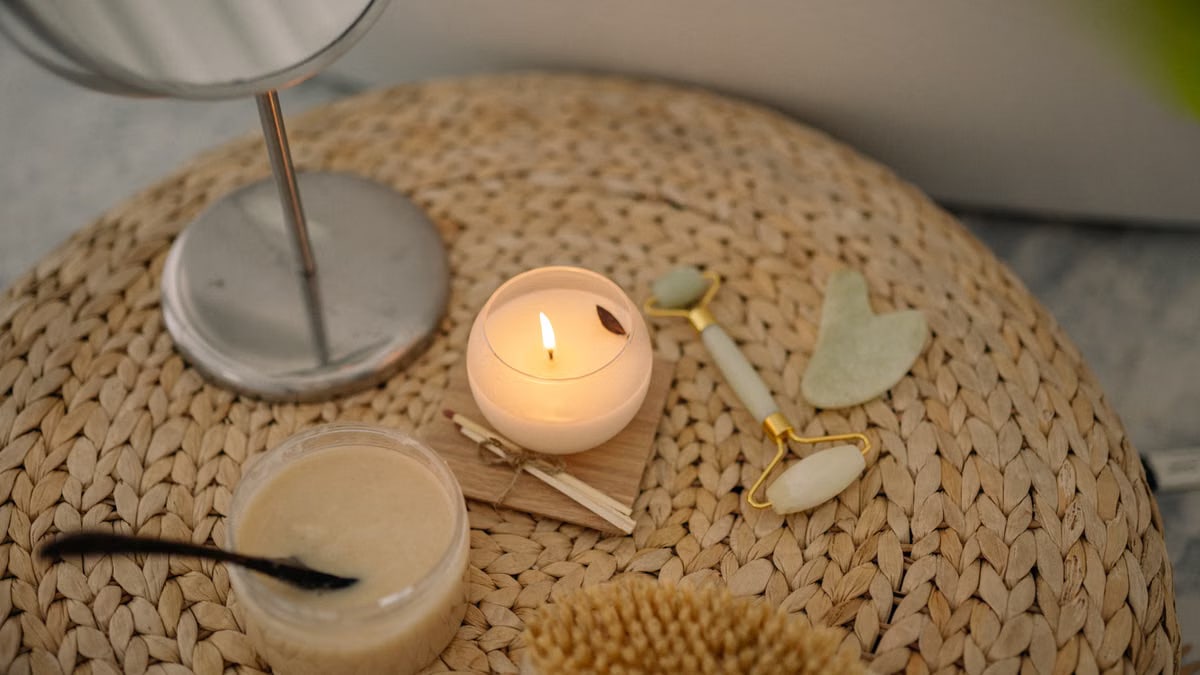 A serene spa setup featuring a lit candle, jade face roller, and body scrub on a woven mat.