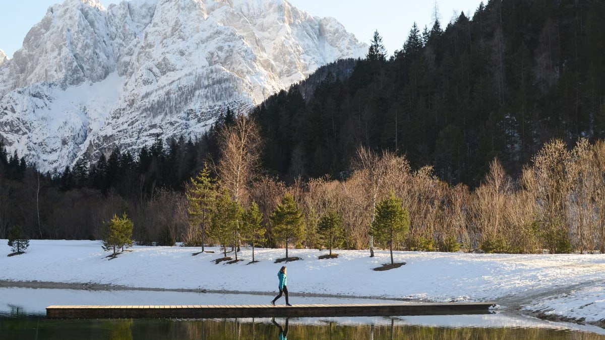 Woman walking on a snowy pier in Kranjska Gora, Slovenia, surrounded by snowcapped mountains and evergreen trees.